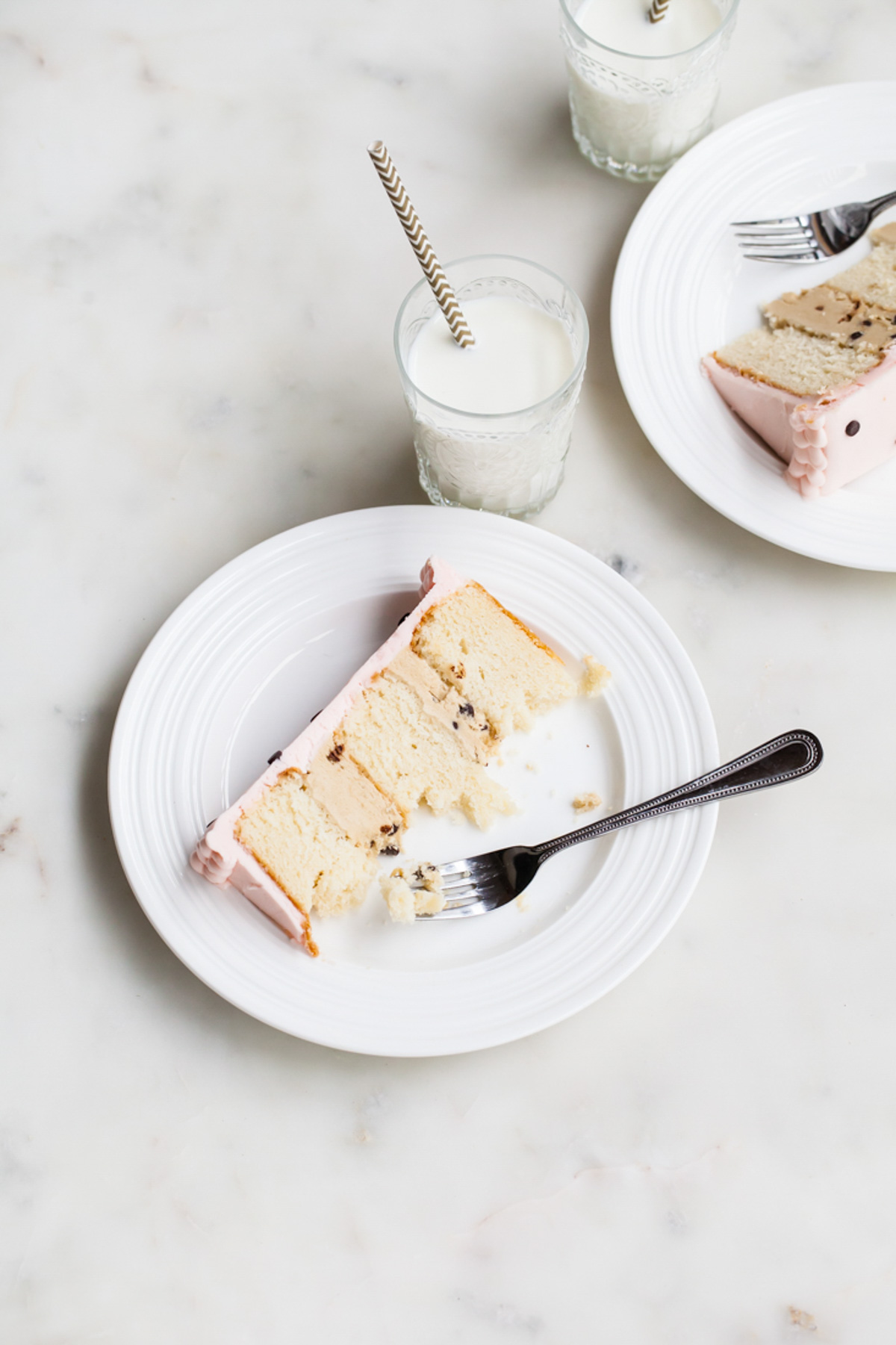 A slice of three-layer cake with pink frosting and cookie dough filling on a plate with a glass of milk