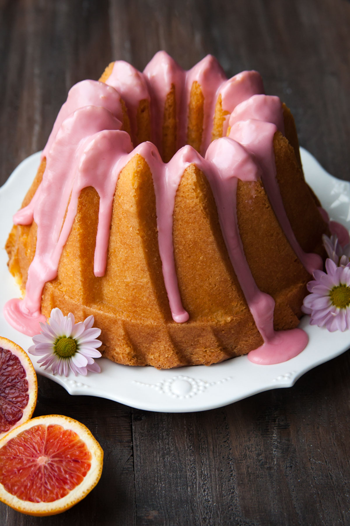 An orange bundt cake on a platter with drips of pink blood orange glaze on top.