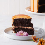 A slice of chocolate caramel cake on a plate with the two layer cake in the background.