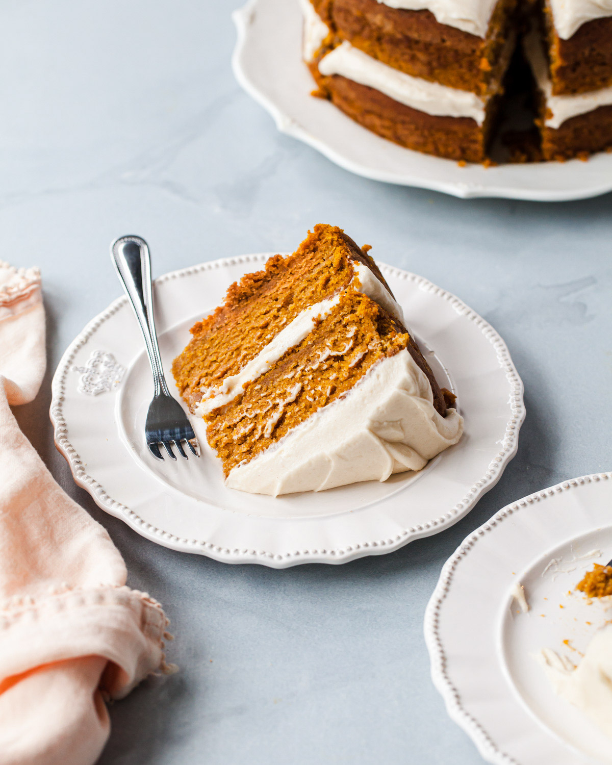 A slice of pumpkin cake with cream cheese frosting being served on a white plate