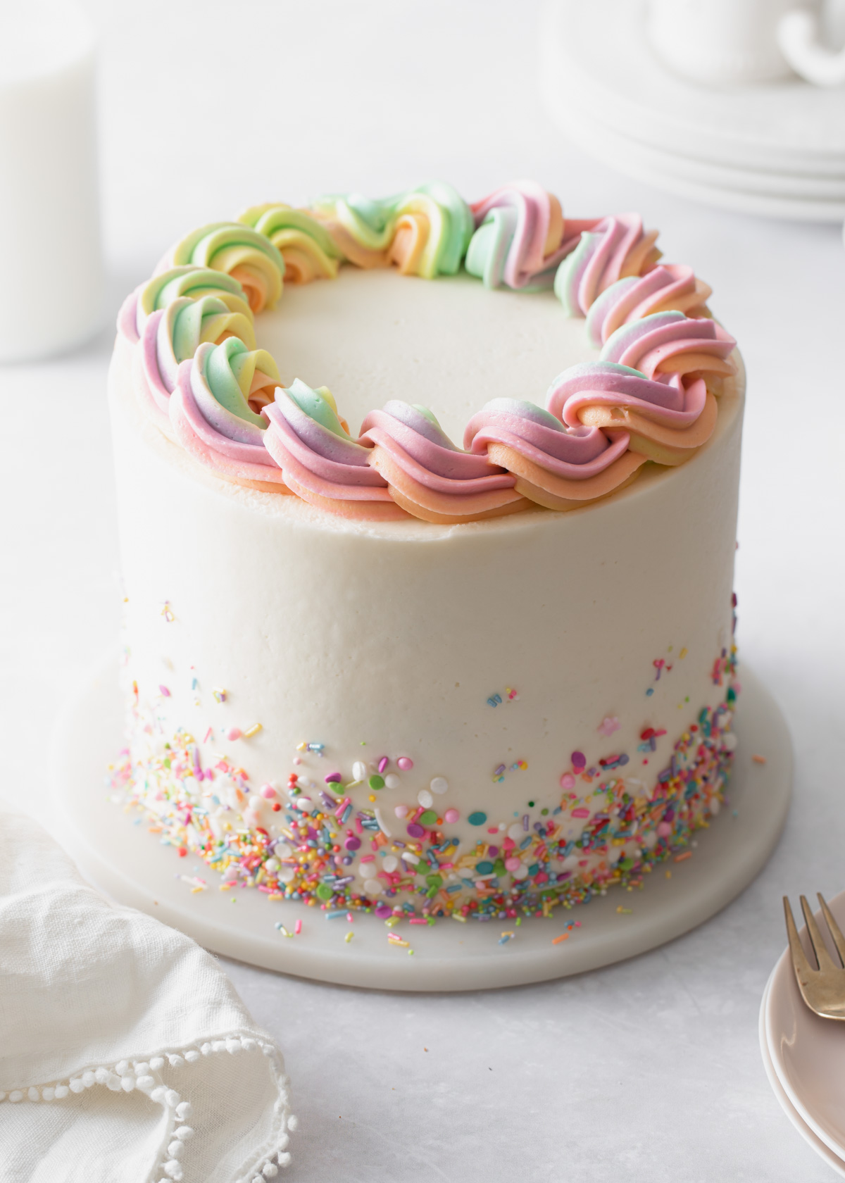 A three-layer sprinkle cake with buttercream frosting and sprinkles scattered around the bottom and rainbow buttercream piped on top