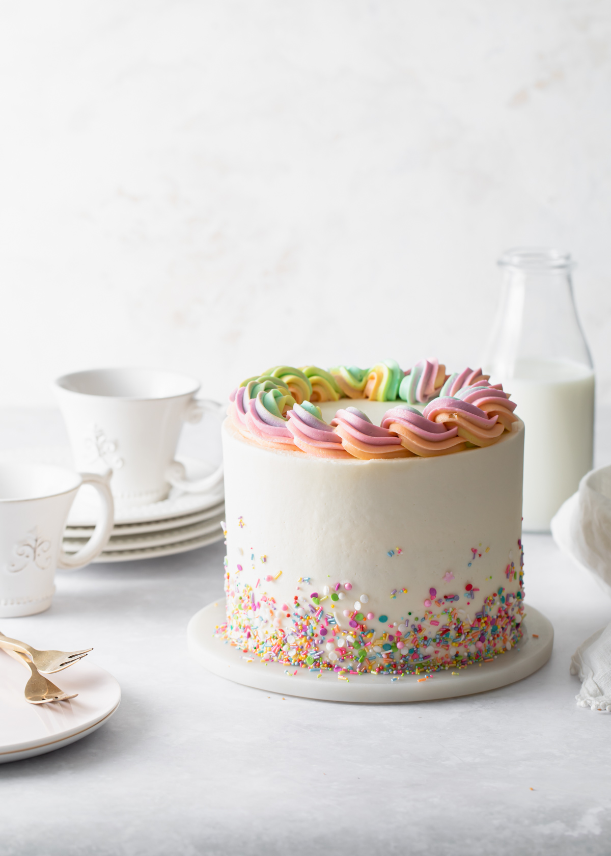 A three-layer sprinkle cake with buttercream frosting and sprinkles scattered around the bottom and rainbow buttercream piped on top
