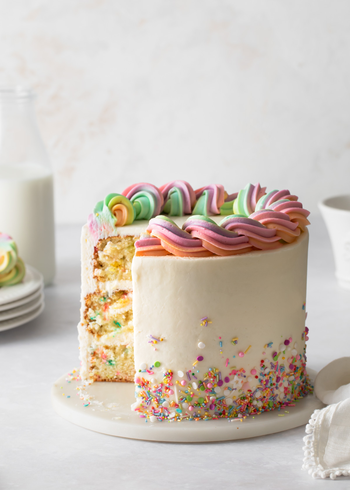 A three-layer sprinkle cake with buttercream frosting and sprinkles scattered around the bottom and rainbow buttercream piped on top