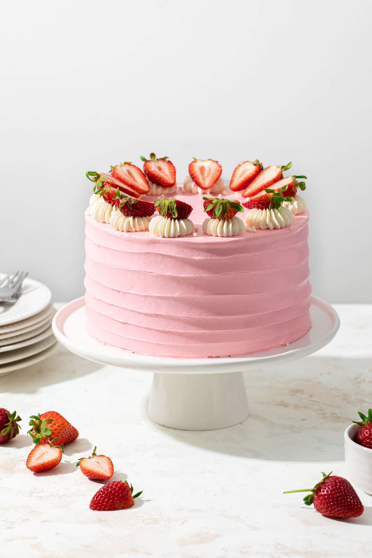 A pink strawberry layer cake on a cake pedestal with fresh strawberries on top