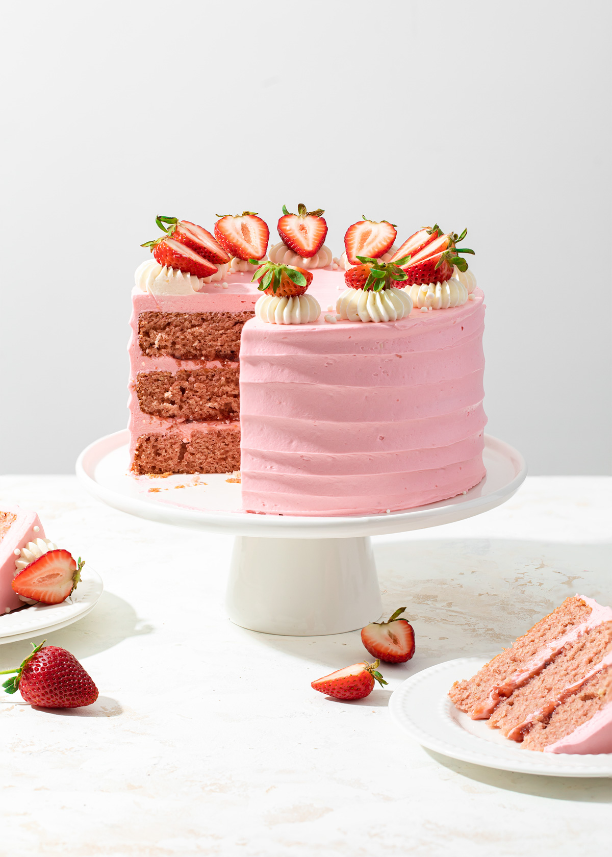 A three-layer strawberry cake with strawberry jam filling and pink frosting on a white cake stand.
