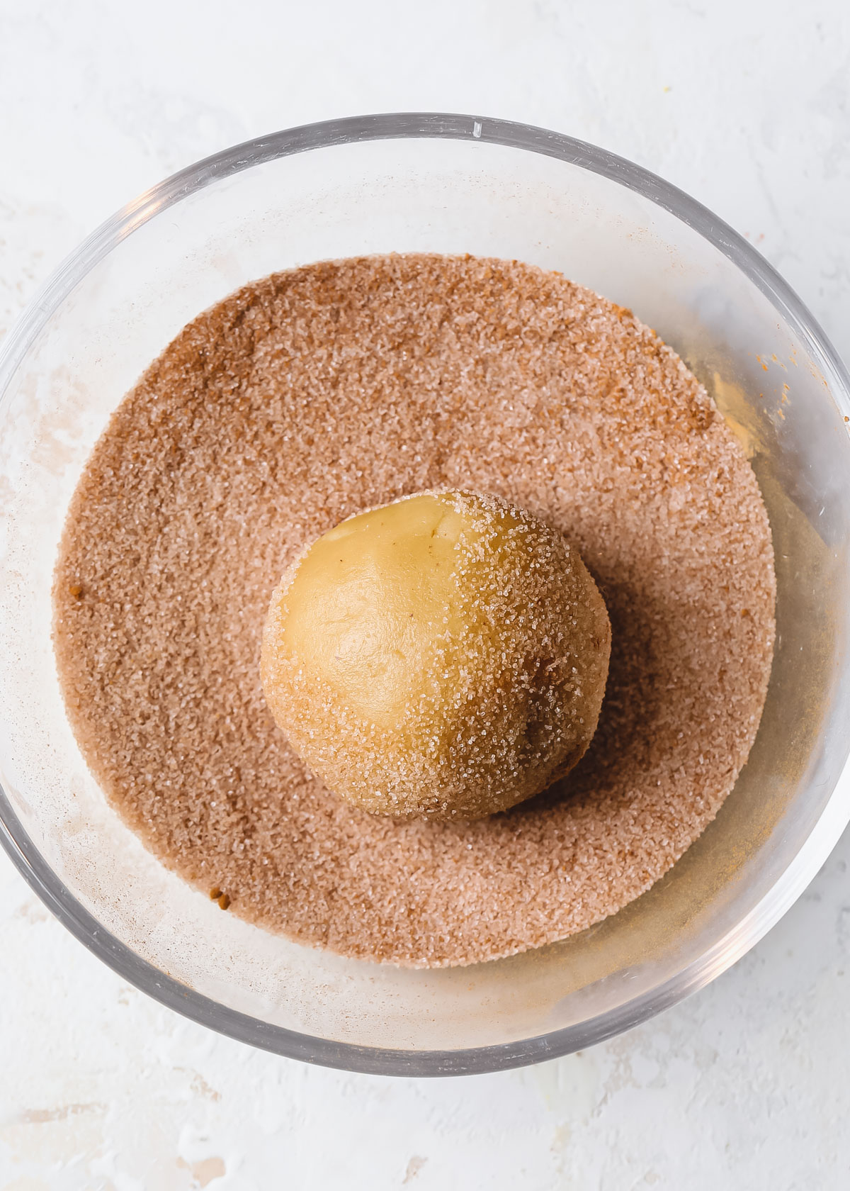 A ball of snickerdoodle cookie dough rolling in a bowl of cinnamon and sugar