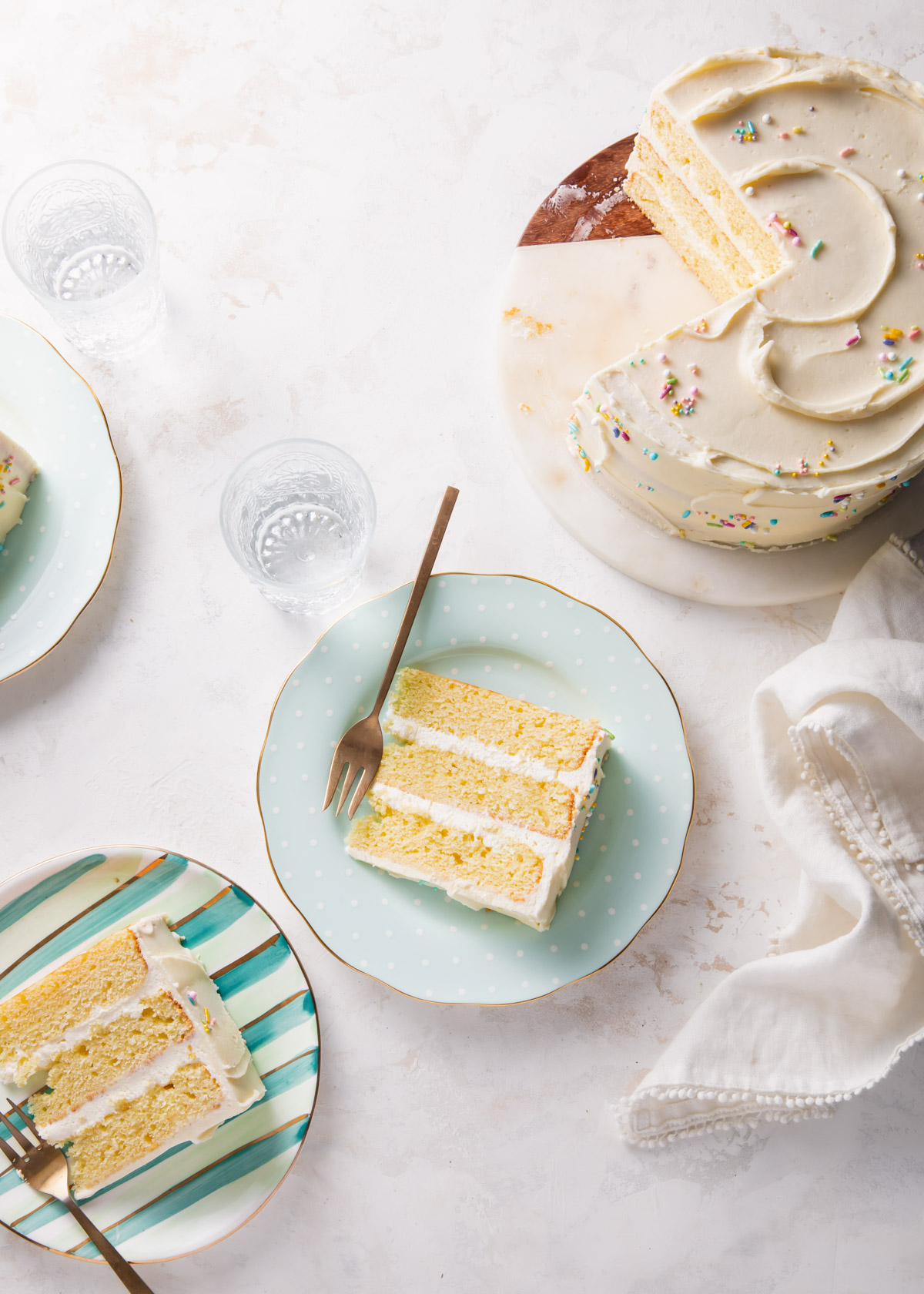 A table with slices of yellow cake on teal plates and an 8 inch cake with buttercream and sprinkles