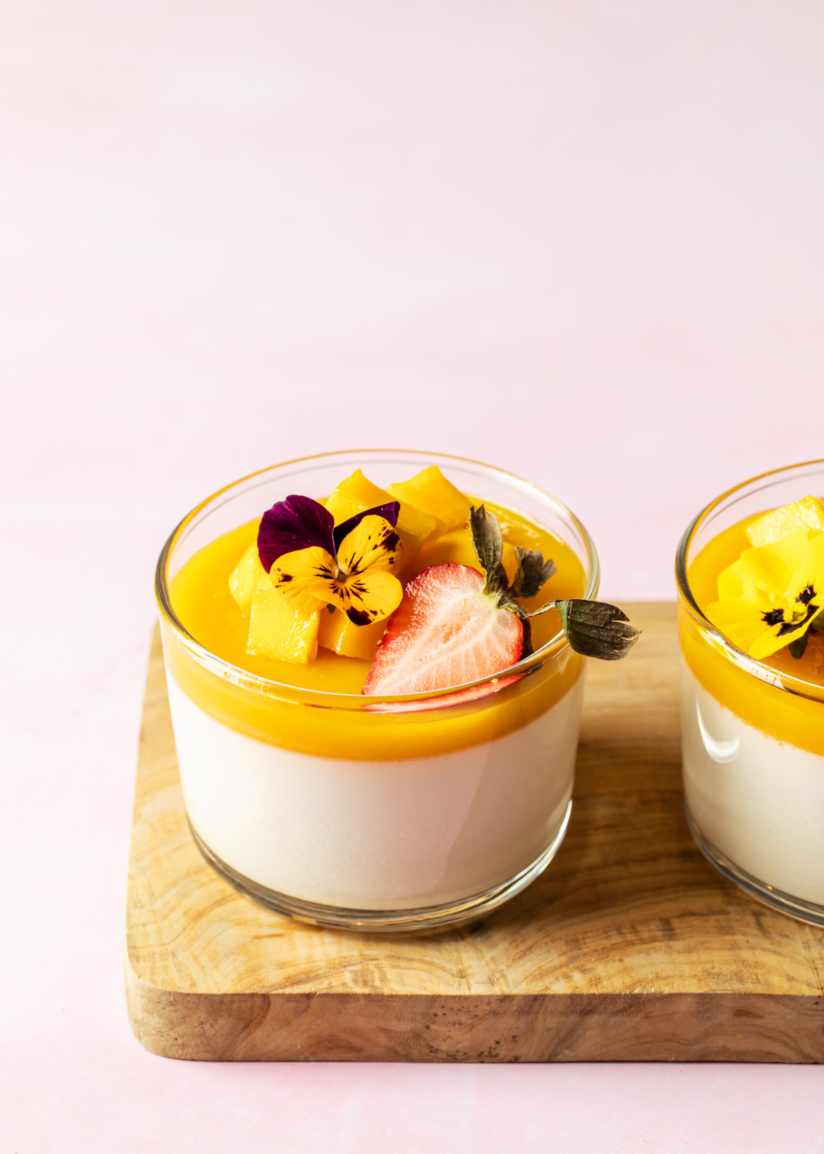 A glass cup of coconut mango panna cotta with fresh fruit and an edible flower on top