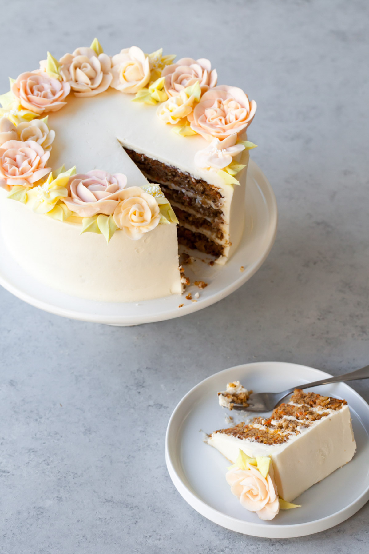 A four layer carrot cake with cream cheese frosting and beautiful buttercream roses on the top with a slice cut out.