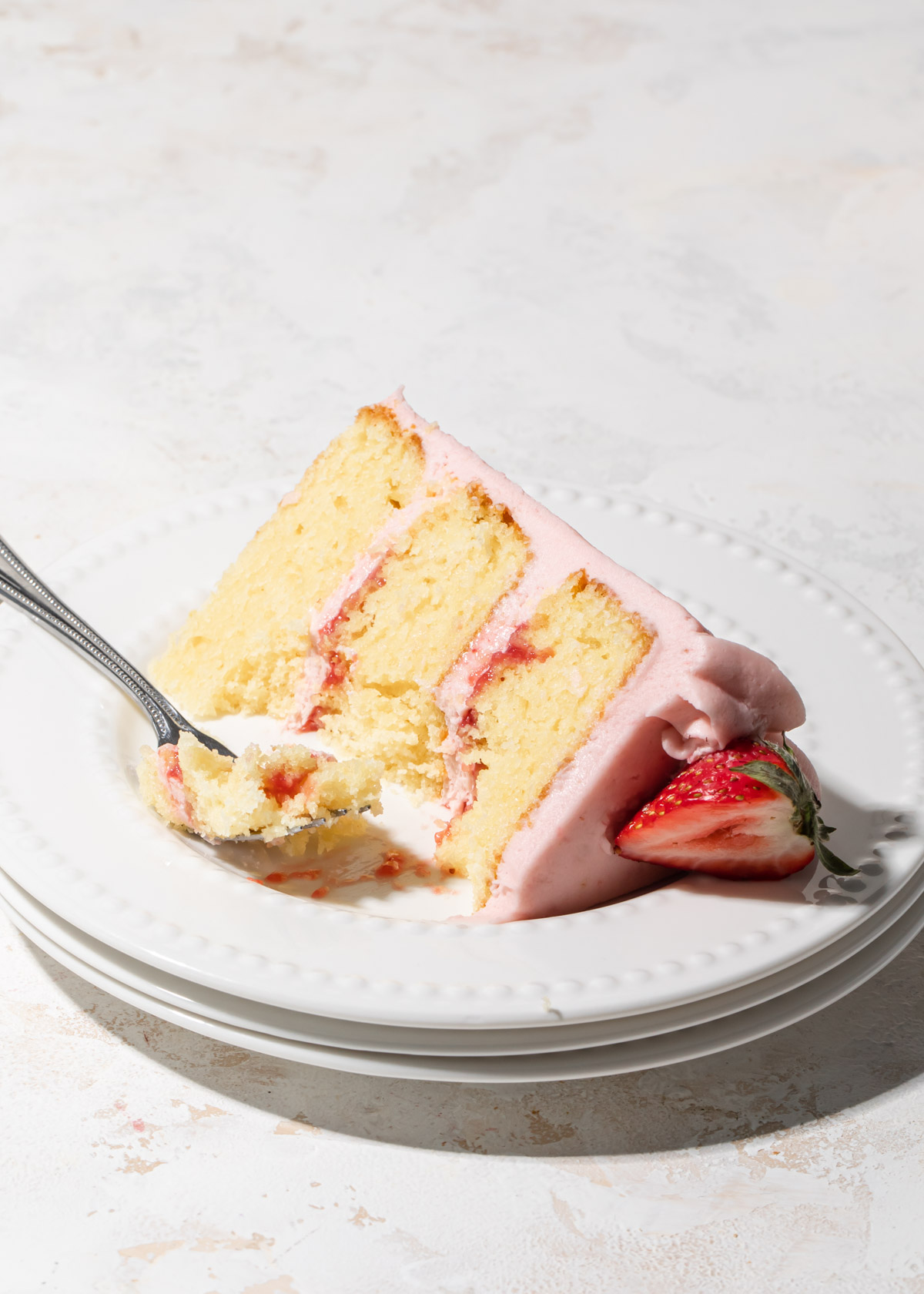 A slice of vanilla cake with strawberry filling and frosting on a stack of white plates
