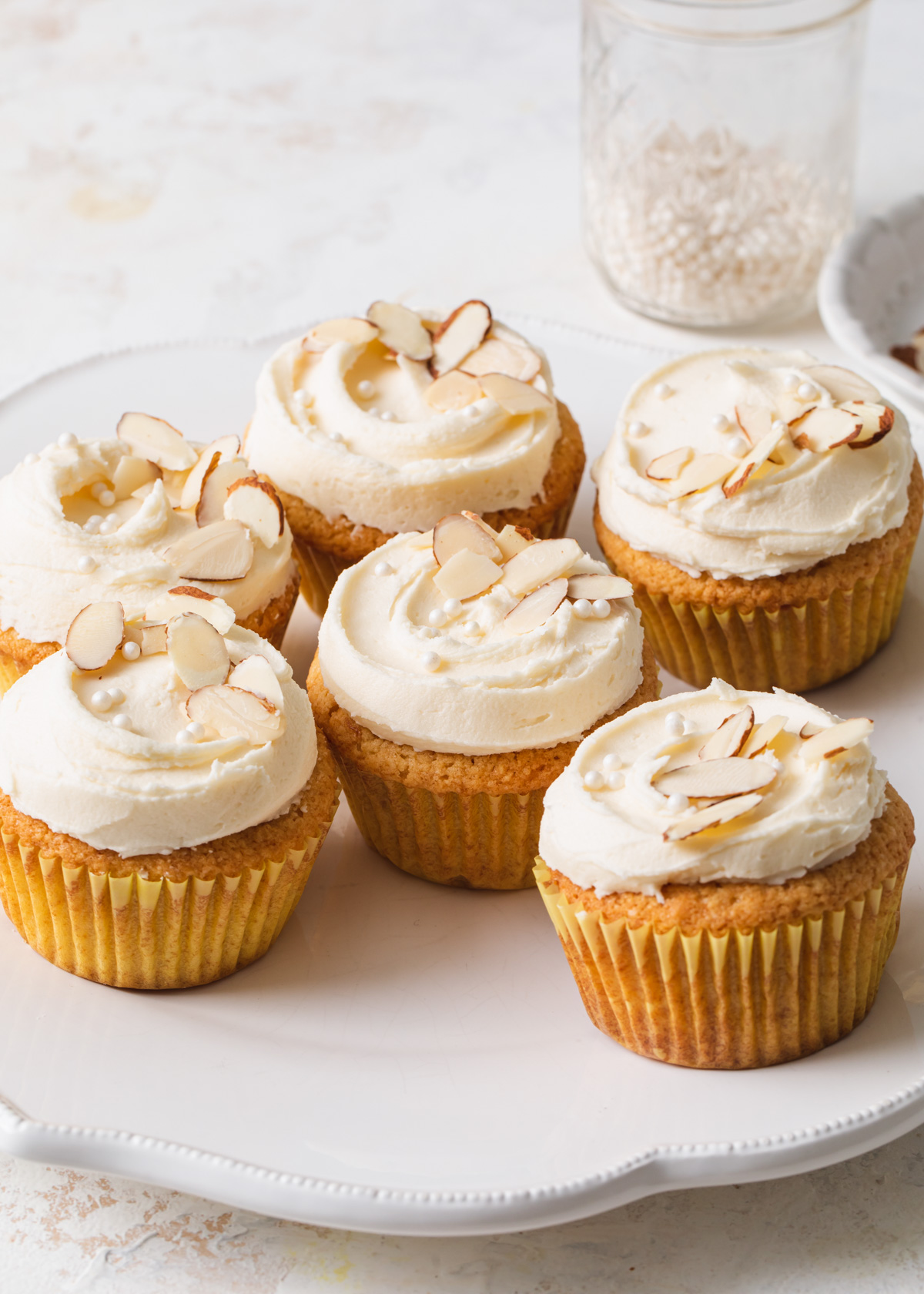 A white platter of almond cupcakes with sliced almonds and sugar pearls on top