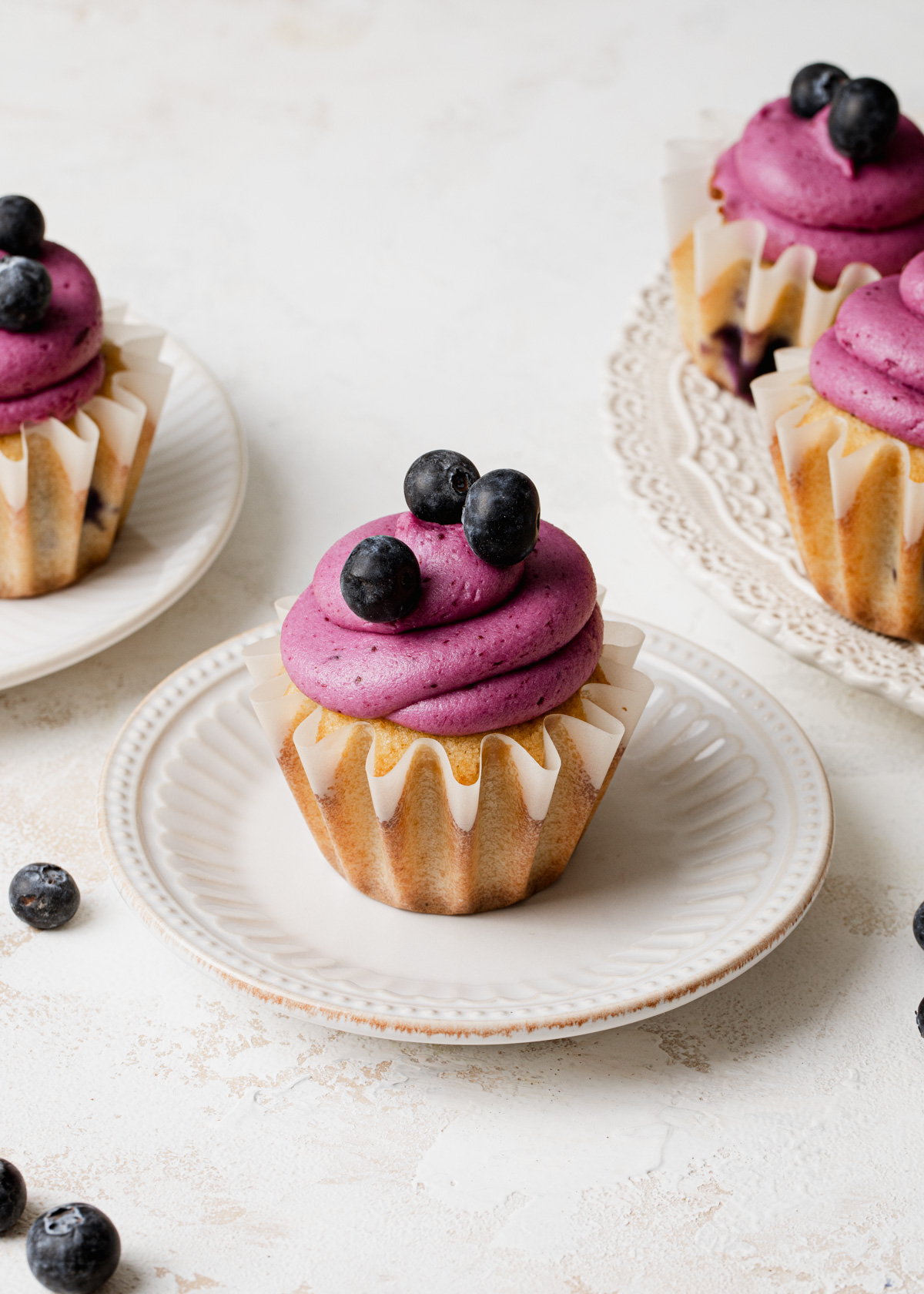 A white plate with a cupcake and purple blueberry buttercream swirled on top and fresh blueberries