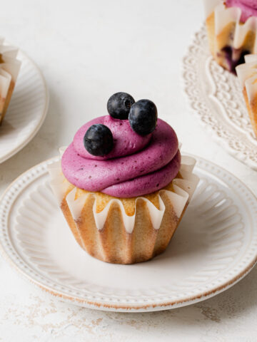A cupcake with purple blueberry frosting swirled on top with a fresh blueberry