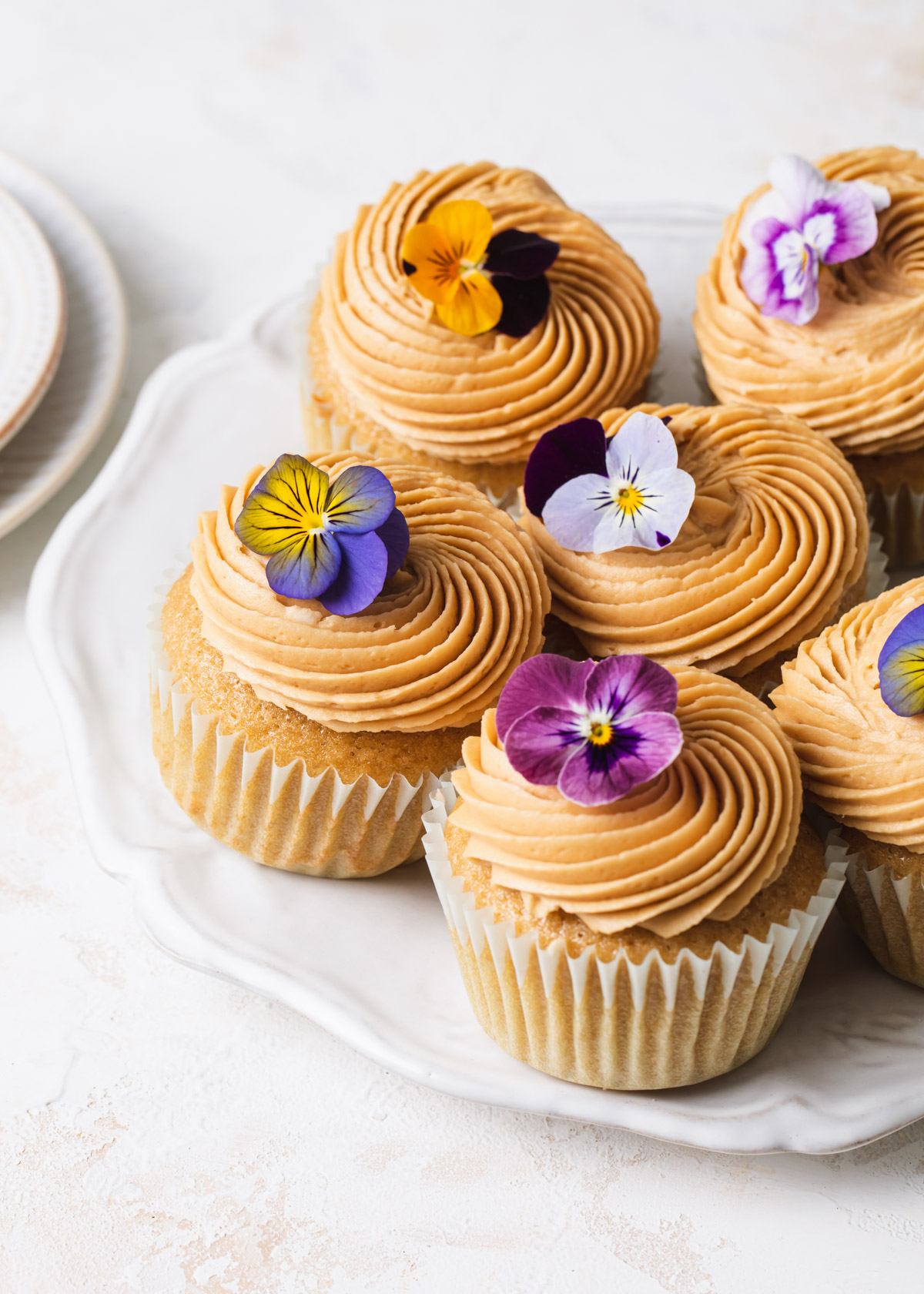 A white background with a plate of coffee cupcakes with edible flowers on top