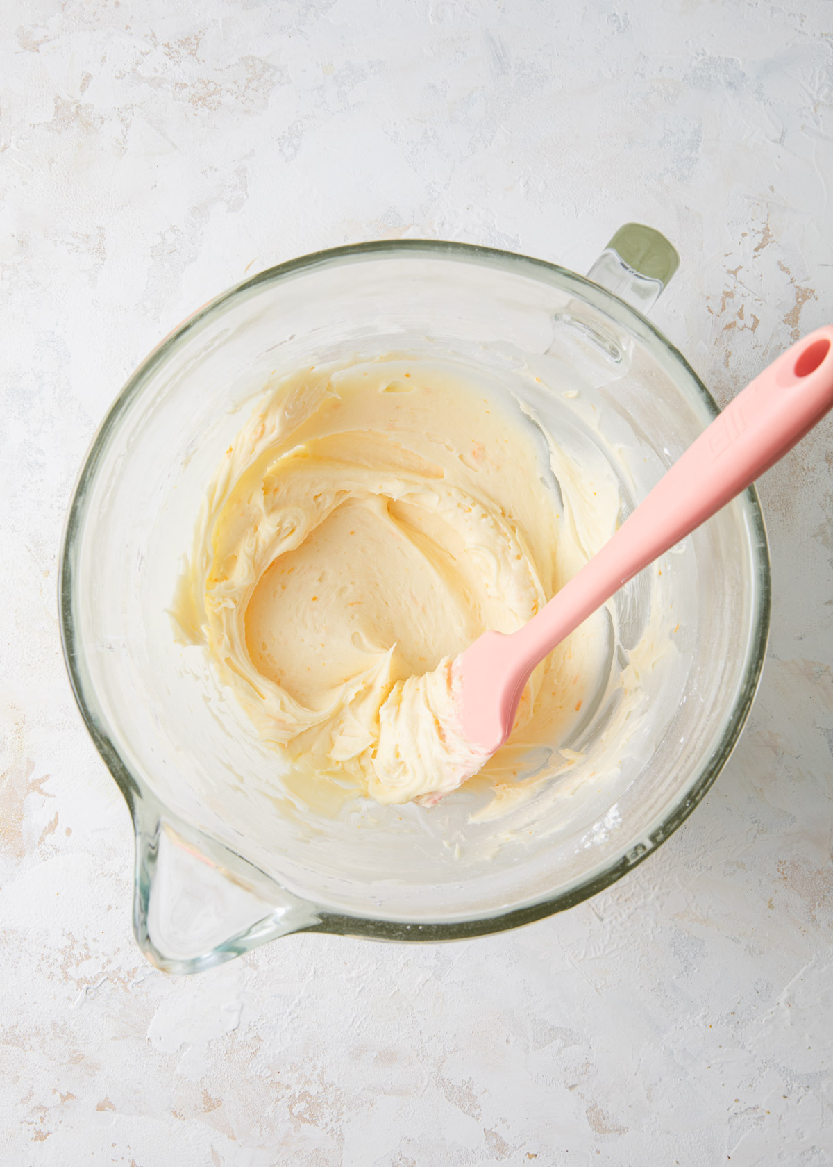 Smooth orange buttercream in a large glass mixing bowl and a spatula