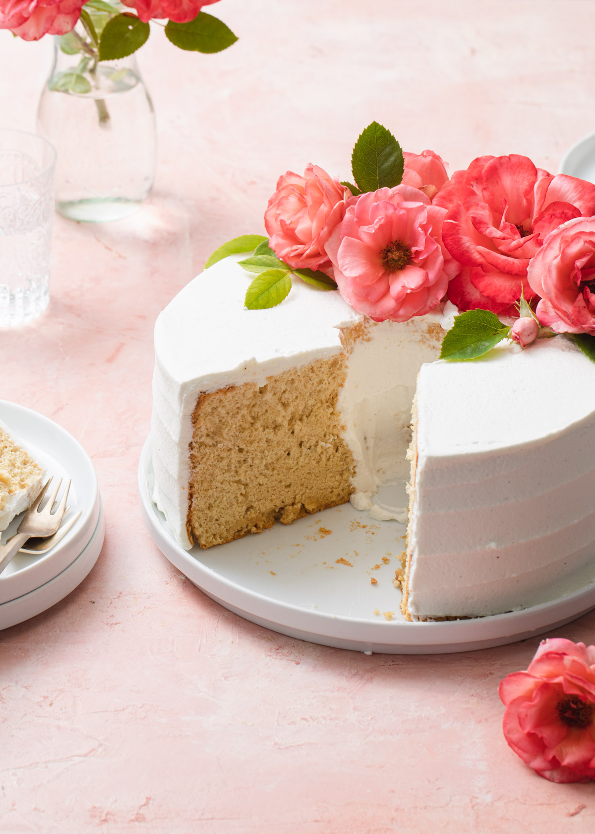 A chiffon cake with whipped cream frosting and fresh pink roses on top set on a white plate