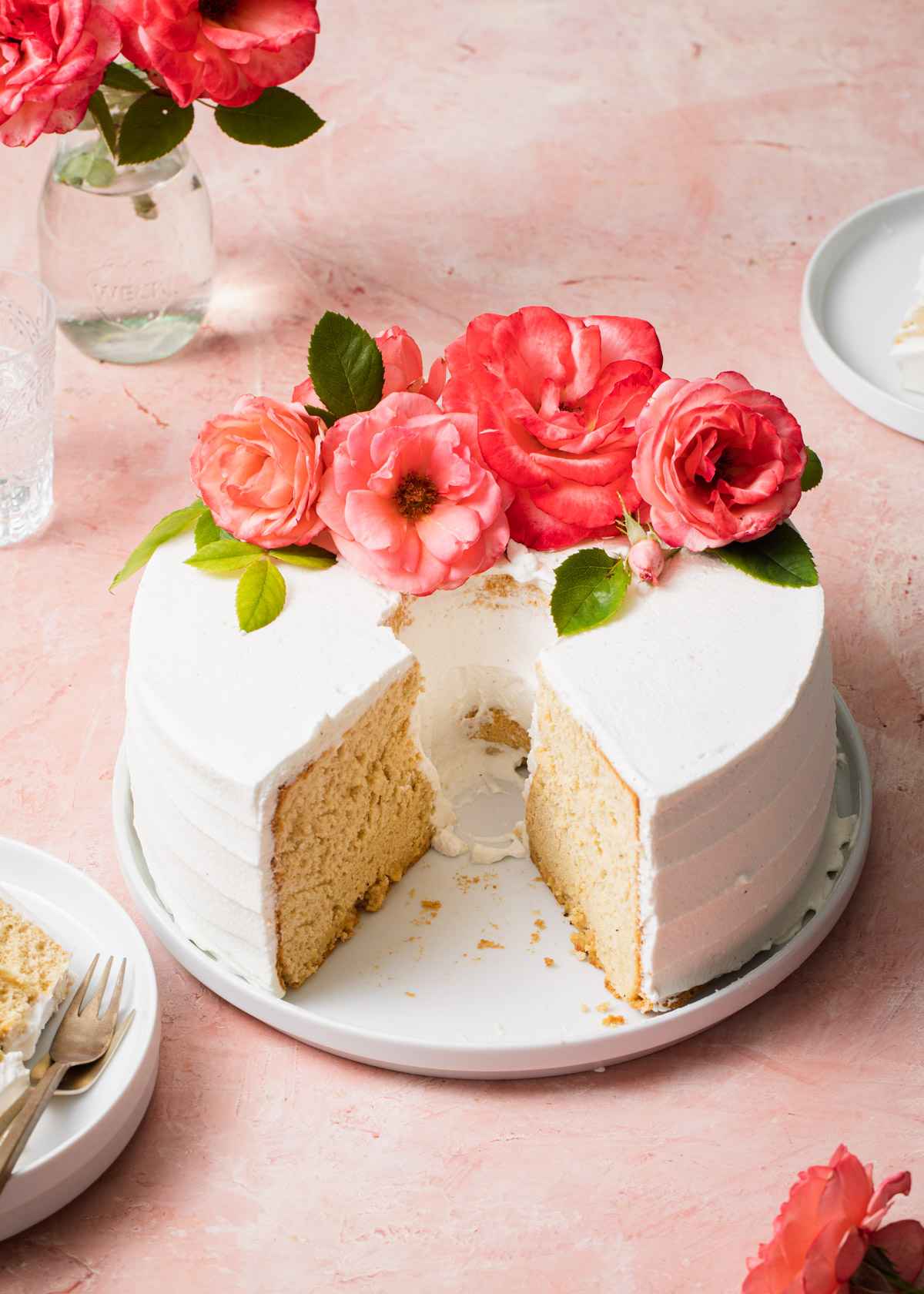 A chiffon cake with whipped cream frosting and fresh pink roses on top set on a white plate