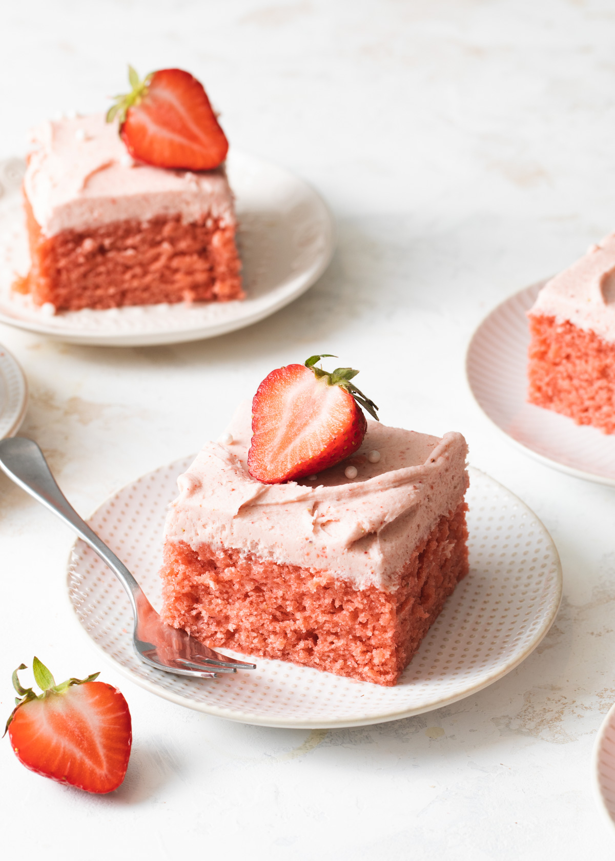A square slice of strawberry cake with strawberry frosting on top of a white plate