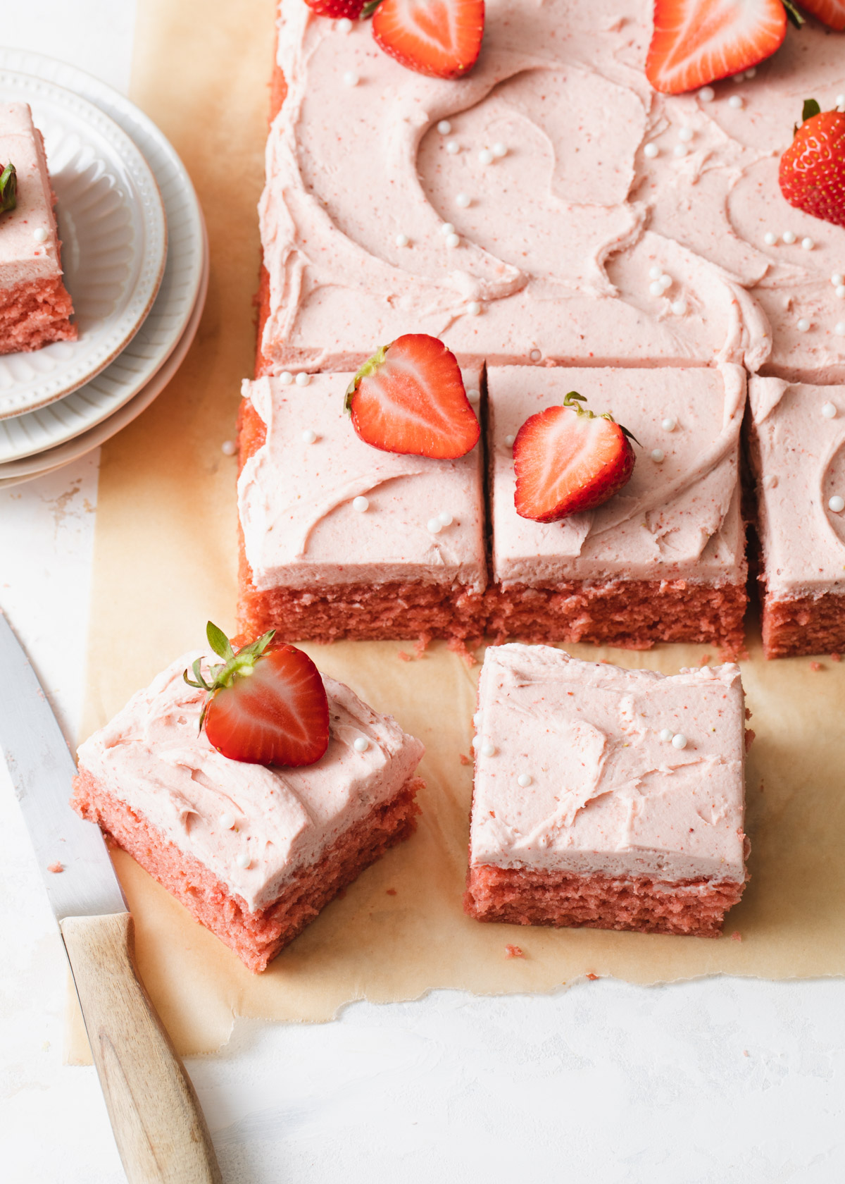 Squares of strawberry sheet cake being cut up on a piece of parchment paper