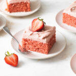 A square slice of strawberry cake with strawberry frosting on top of a white plate