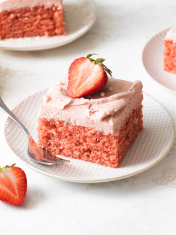 A square slice of strawberry cake with strawberry frosting on top of a white plate