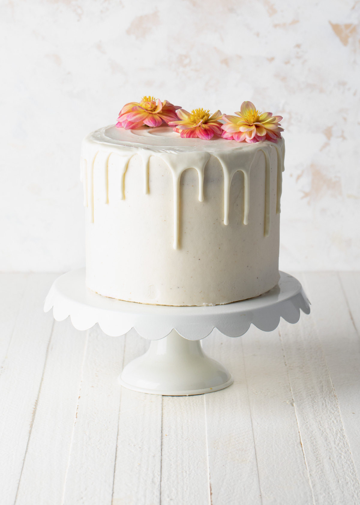 An all white cake with a white chocolate drip on a white cake stand.