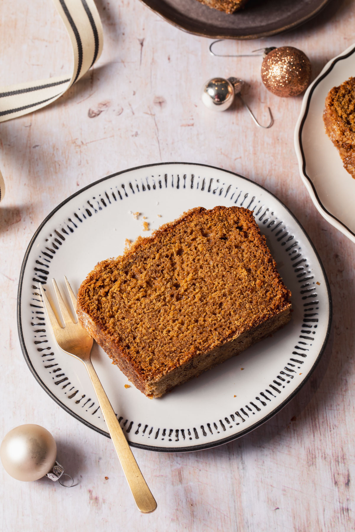 A slice of ginger loaf cake on a plate set on a wooden table with a gold fork
