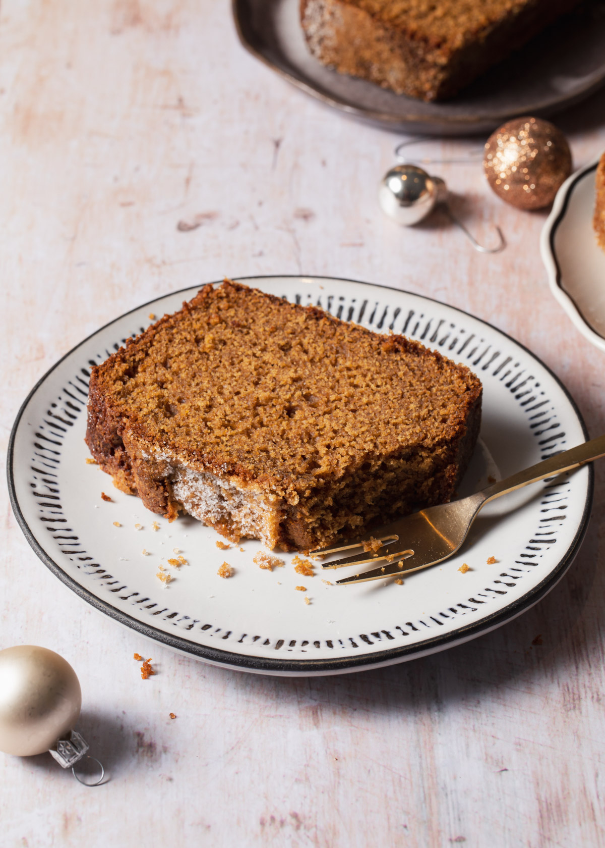 A slice of moist gingerbread cake on a white plate with metallic Christmas ornaments in the background 