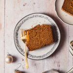 A thick slice of gingerbread loaf cake on a white plate set on a wooden tabletop