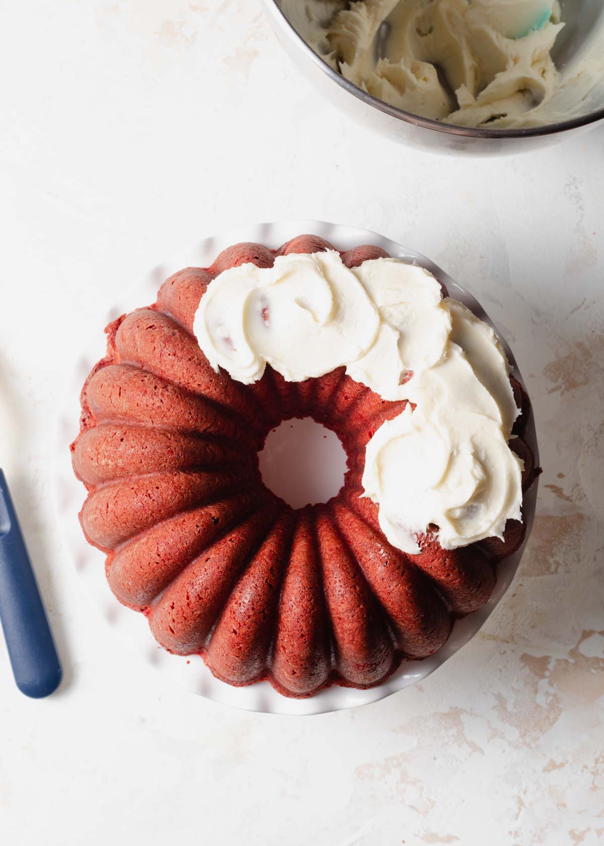 A red velvet Bundt Cake on a white cake stand with fluffy cream cheese frosting