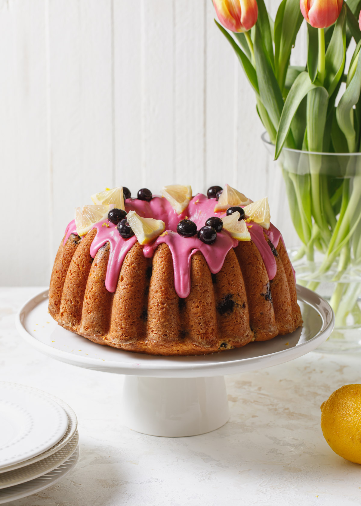 A lemon blueberry bundt cake with blueberry icing and decorated with lemon slices on a white cake stand with flowers in the background