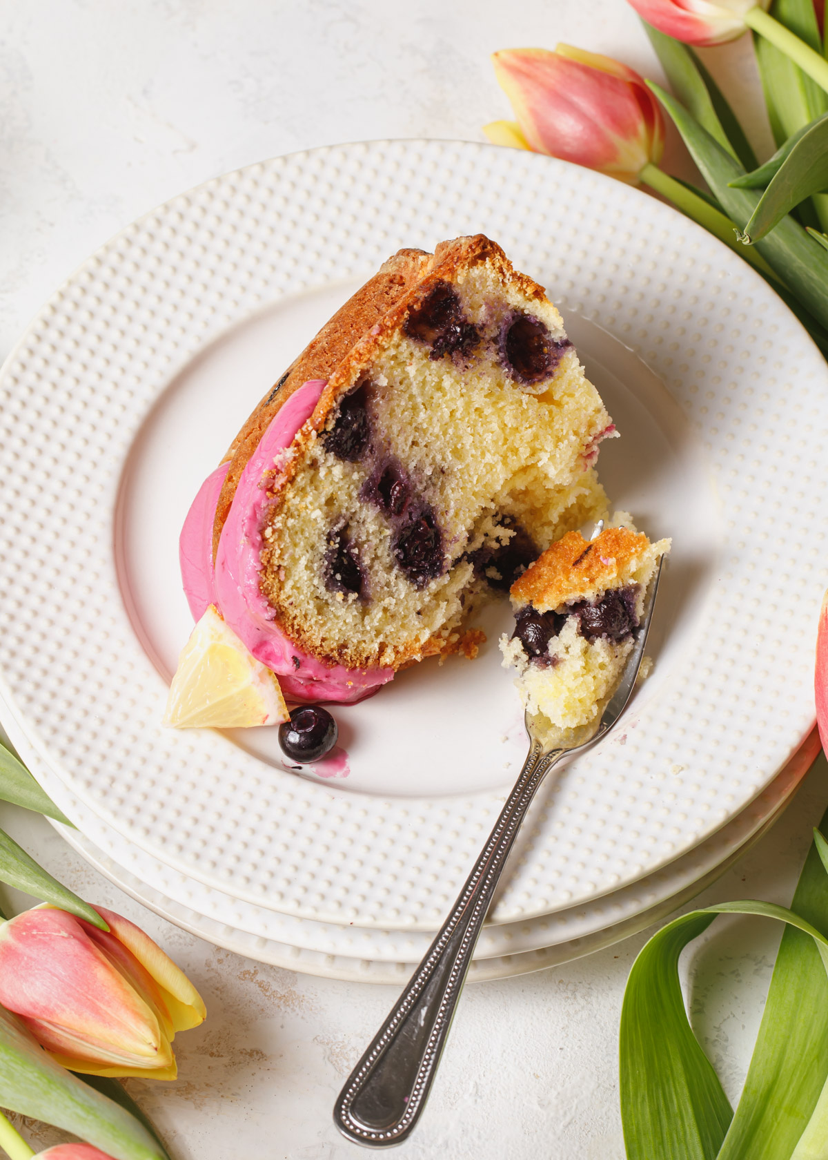 A slice of lemon blueberry pound cake on a white plate with flowers in the background