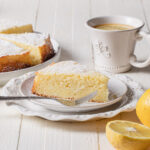 A thick slice of lemon ricotta cake with lemons and tea in the background