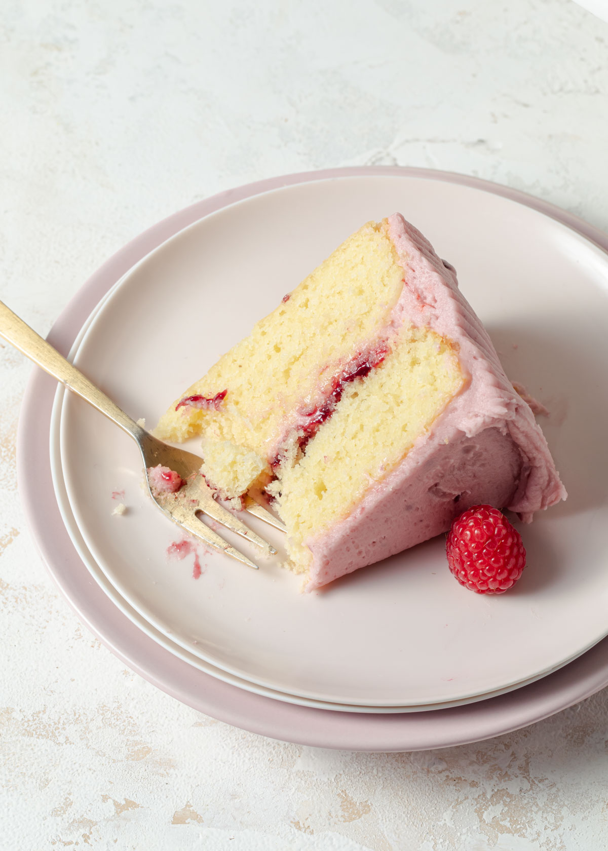 A slice of almond layer cake with raspberry filling and frosting set on a pale pink plate.