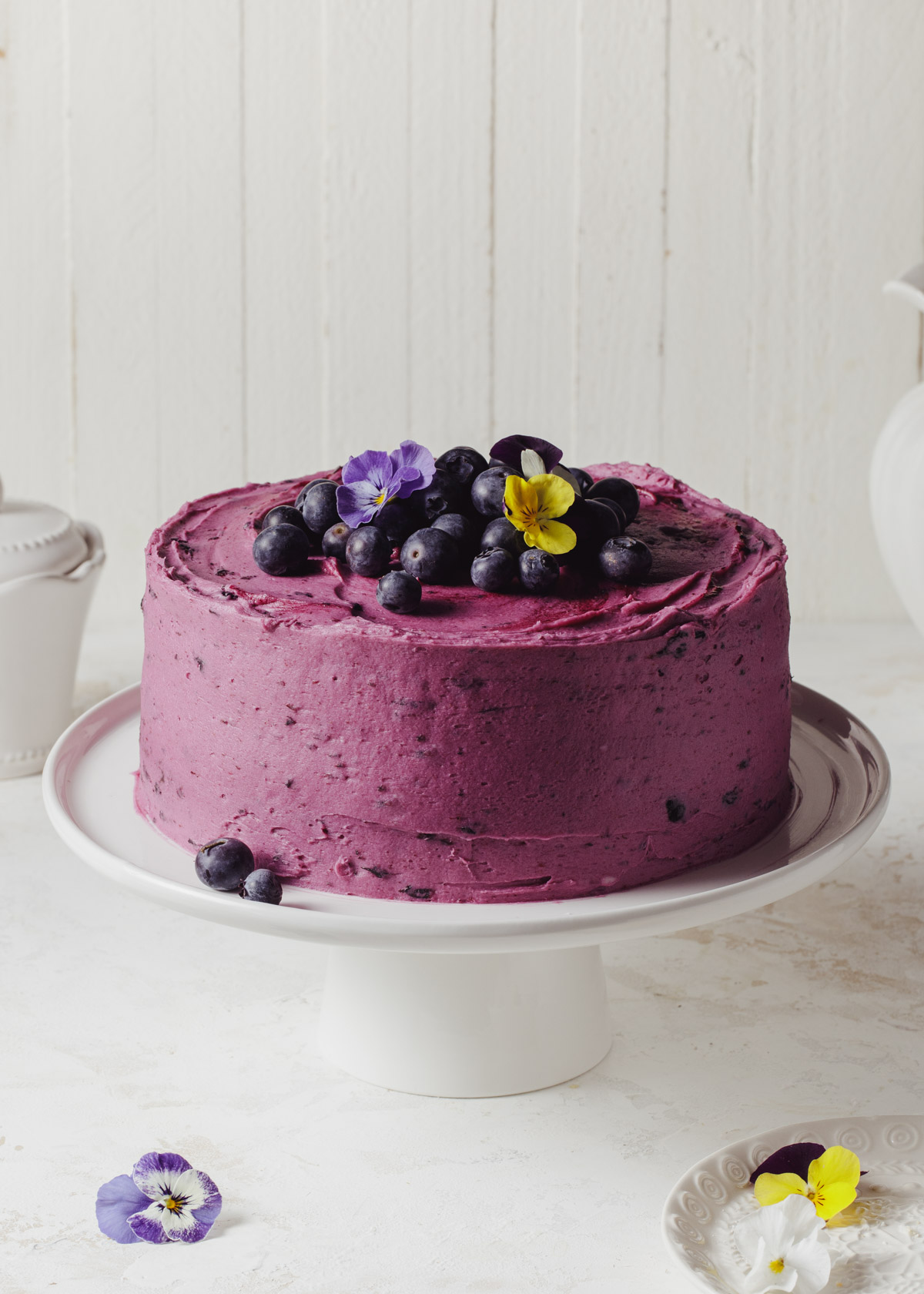 A purple buttercream blueberry chocolate cake with fresh blueberries on top set on a white cake stand