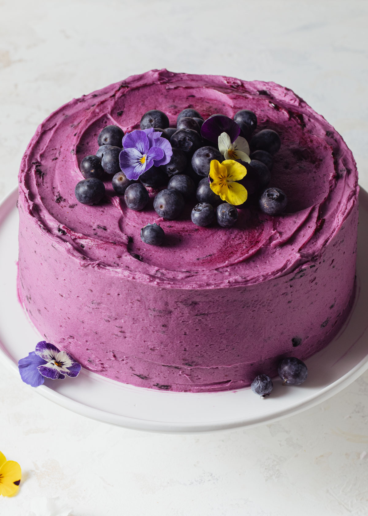 A purple buttercream blueberry chocolate cake with fresh blueberries on top set on a white background